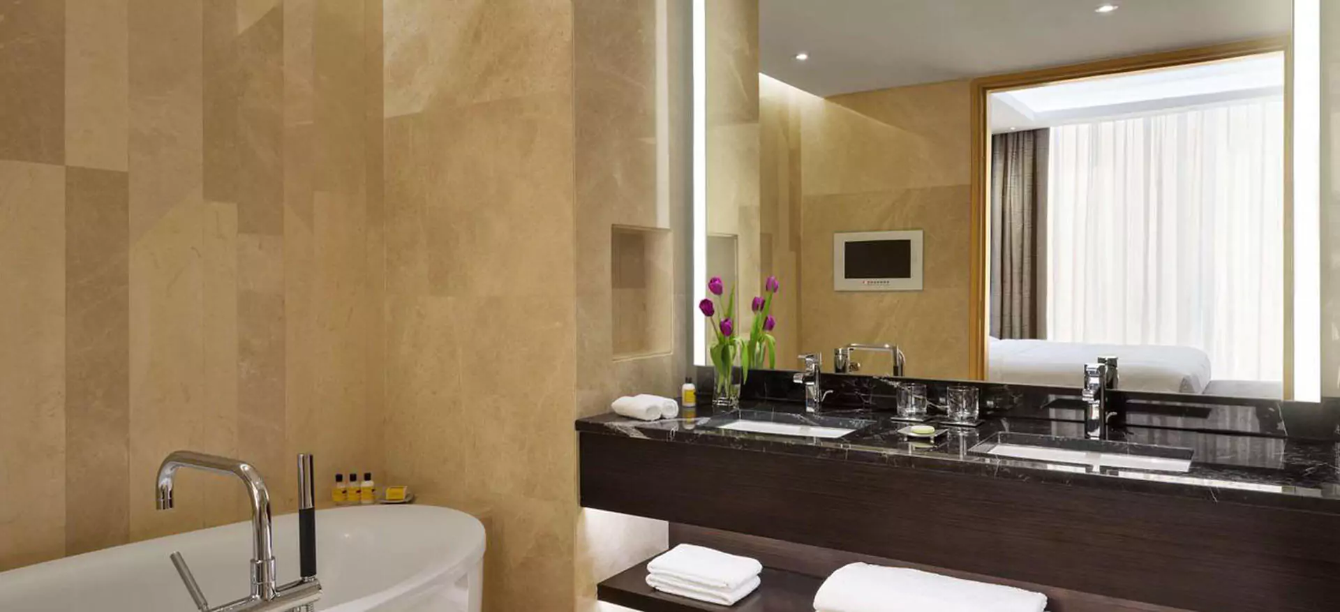 Bathroom with a bathtub and a lighted mirror above two sinks on a black marble countertop in Oman.